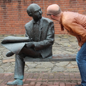 Ein Mann in einer braunen Jacke beugt sich vor, um einer Bronzestatue eines sitzenden, bärtigen Mannes, der eine Zeitung liest, etwas zuzuflüstern.