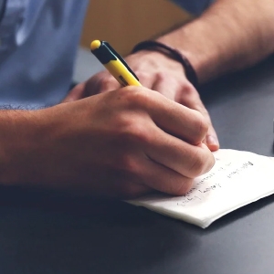 Eine Person macht sich Notizen in einem kleinen Notizbuch auf einem dunklen Schreibtisch und hält einen Stift mit gelber Spitze in der Hand.
