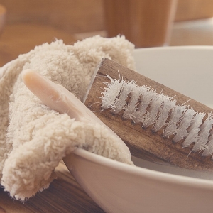 Eine hölzerne Nagelbürste, ein Stück Seife und ein flauschiges beiges Handtuch liegen neben einer weißen Keramikschüssel.