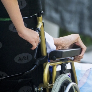 Nahaufnahme der Hand einer älteren Person auf der Armlehne eines Rollstuhls, auf der sanft die Hand einer anderen Person ruht.