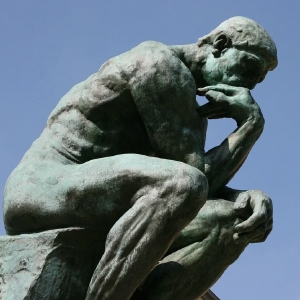 Bronzestatue „Der Denker“ von Rodin. Sie zeigt einen muskulösen, sitzenden Mann, der sein Kinn in tiefer Kontemplation vor einem klaren blauen Himmel auf eine Hand stützt.