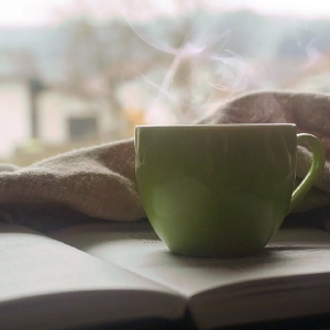 Eine dampfende grüne Tasse auf einem offenen Buch mit einer gemütlichen Decke und einem unscharfen Fensterhintergrund.