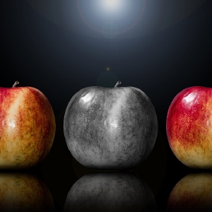 Drei Äpfel, einer rot, einer grau und einer rot-gelb, in einer Reihe unter einem Scheinwerfer auf einem dunklen Hintergrund mit sichtbaren Reflexionen angeordnet.
