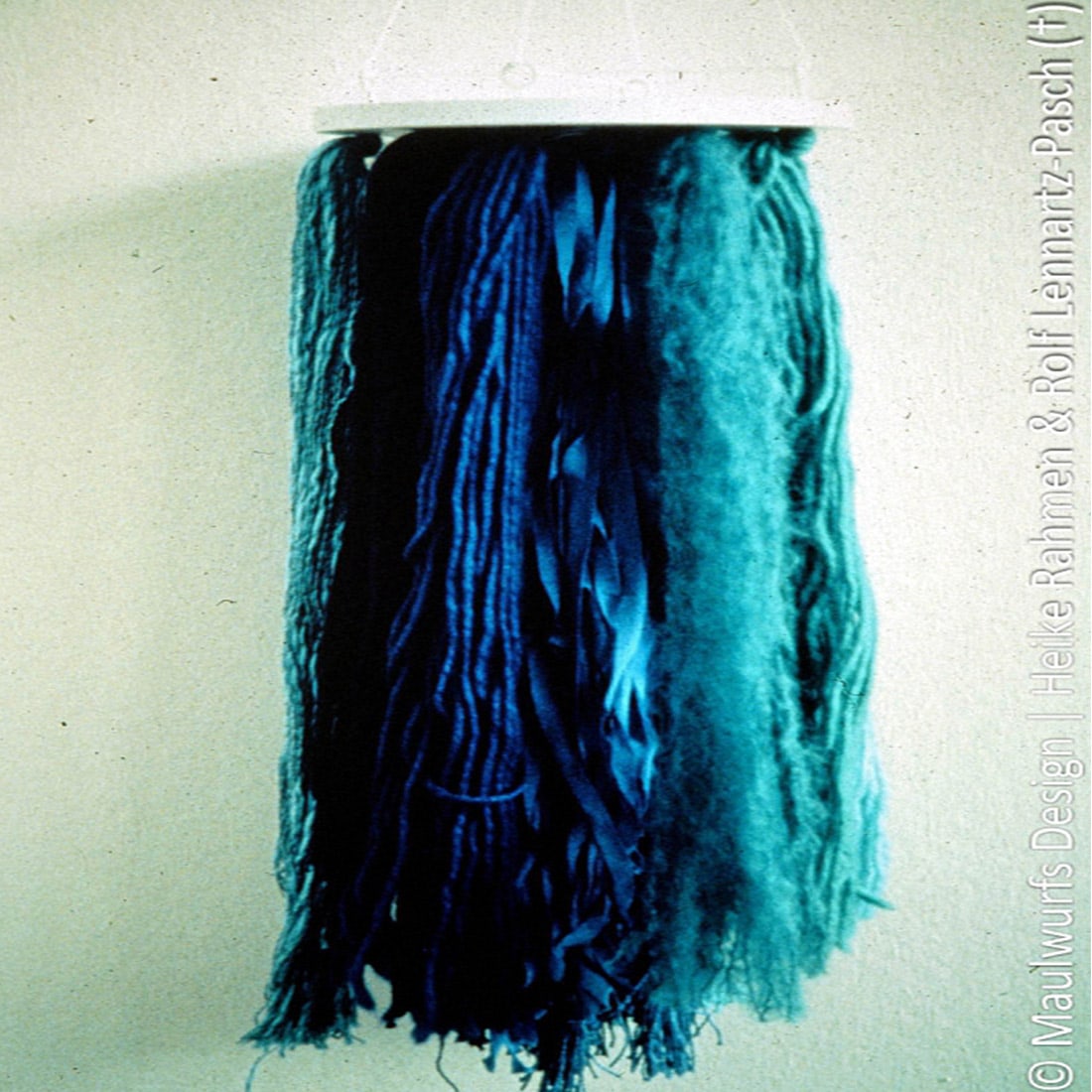 Ein an der Wand montiertes Textilkunstwerk mit langen Garnsträngen in Blautönen, dargestellt in einem Farbverlauf von Hell- zu Dunkelblau.