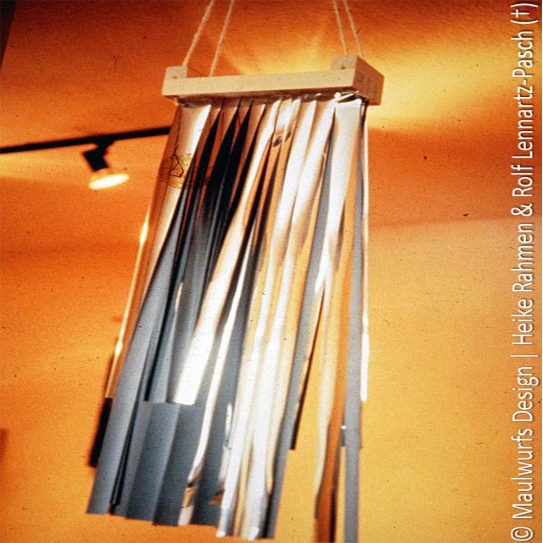Eine Sammlung silberner Metallstäbe hängt vertikal an einem Holzrahmen vor einem orangefarbenen Hintergrund.