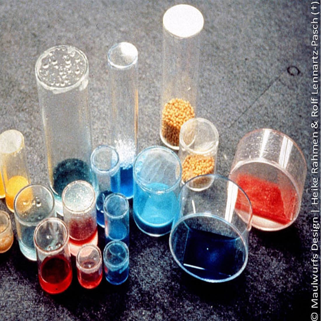 Verschiedene chemische Substanzen in durchsichtigen Laborbehältern in verschiedenen Farben, präsentiert auf einer grauen Oberfläche.