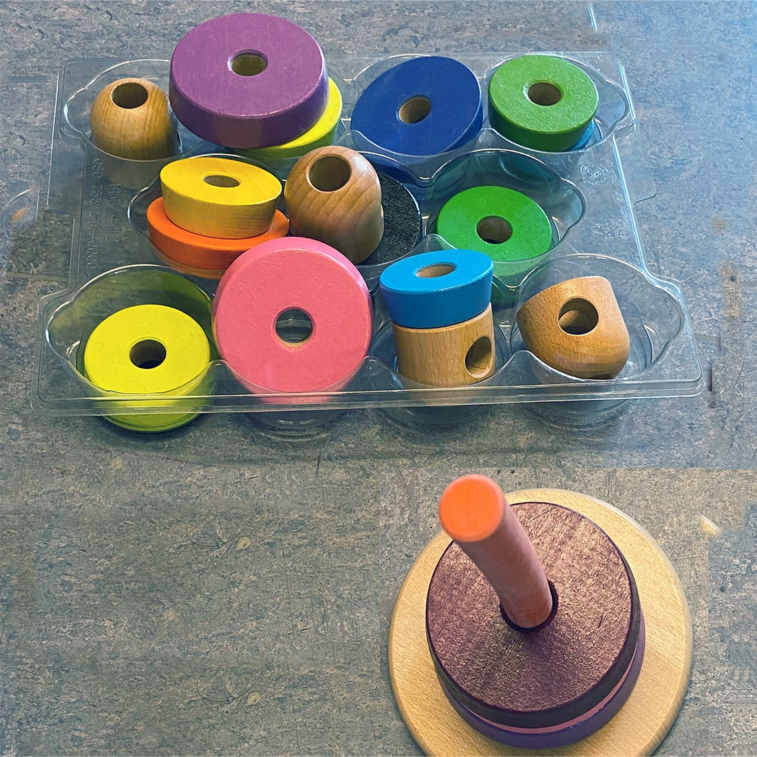 Ein Stapelspielzeug aus Holz mit bunten, abgerundeten Teilen neben seiner Kunststoffverpackung auf einer Tischplatte.