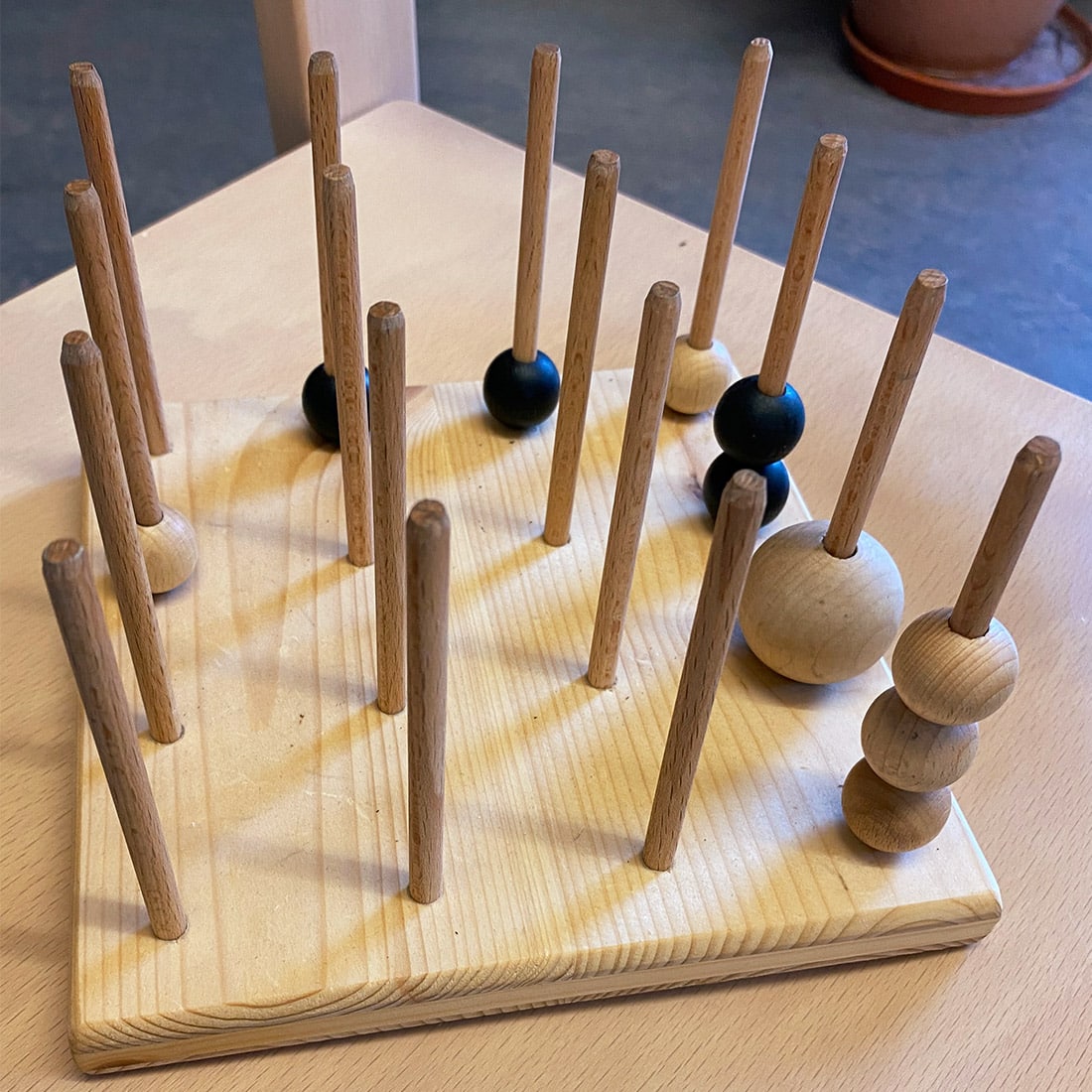 Ein hölzernes Brettspiel mit vertikalen Stiften, von denen einige schwarze und naturfarbene Holzkugeln halten, auf einem Tisch.