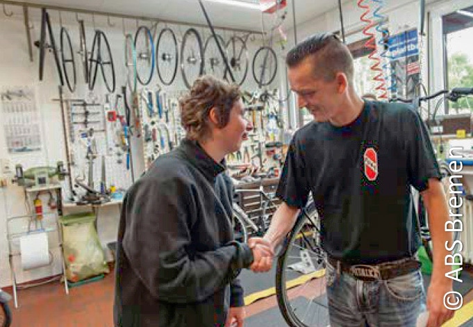 Zwei Menschen geben sich in einer Fahrradwerkstatt die Hand, umgeben von verschiedenen Werkzeugen und Fahrradteilen.