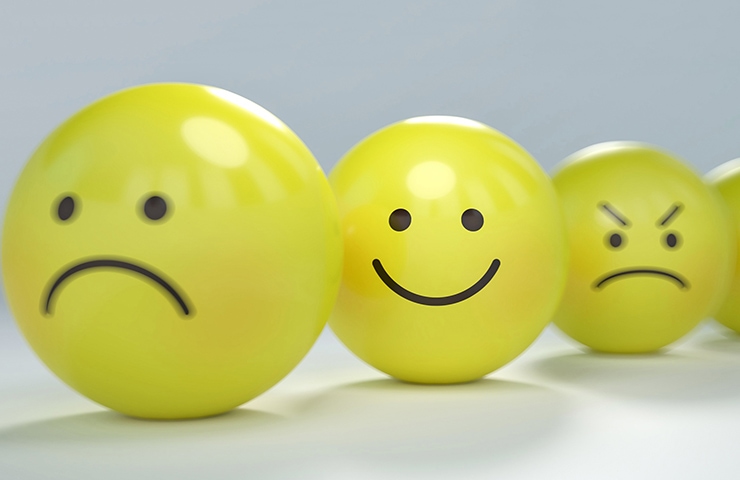 Vier gelbe Emoticon-Bälle zeigen vor einem sanften grauen Hintergrund unterschiedliche Emotionen von Traurigkeit und Wut bis hin zu Freude.