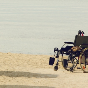 Ein leerer Rollstuhl, der an einem Sandstrand mit ruhigem Wasser im Hintergrund geparkt ist und an dem persönliche Gegenstände hängen.
