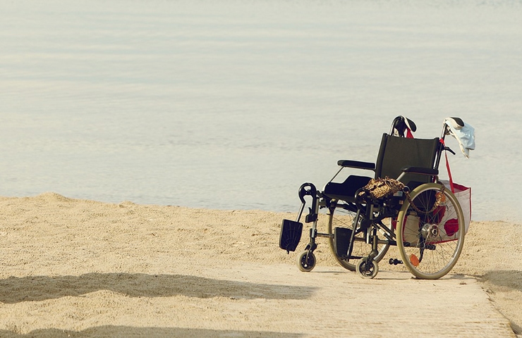 Ein leerer Rollstuhl, der an einem Sandstrand mit ruhigem Wasser im Hintergrund geparkt ist und an dem persönliche Gegenstände hängen.