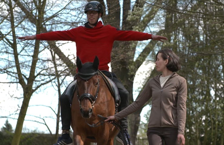 Eine Person in einem roten Pullover balanciert mit ausgestreckten Armen auf einem Pferd, gelenkt von einer anderen Person zu Fuß in einem braunen Oberteil, in einer Waldlandschaft.