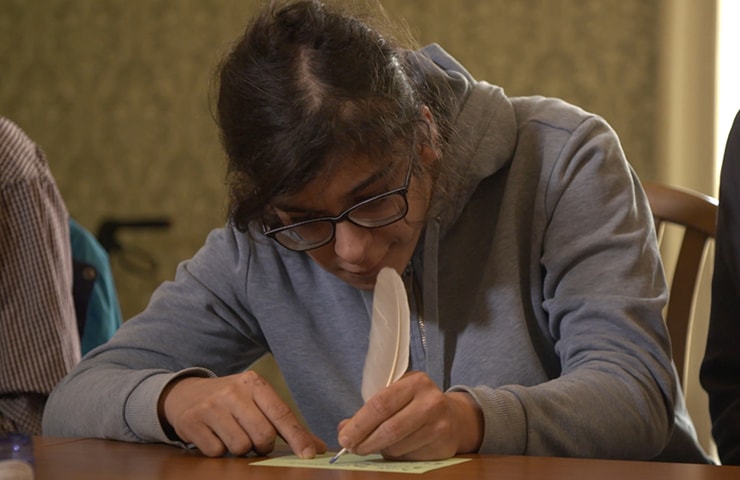 Eine junge Frau mit Brille beugt sich über einen Tisch und schreibt konzentriert eine Notiz mit einem Stift in einem hell erleuchteten Raum.
