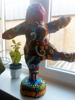 Farbenfroh bemalte Statue einer Figur in dynamischer Pose, neben einem Fenster neben einer Topfpflanze platziert.