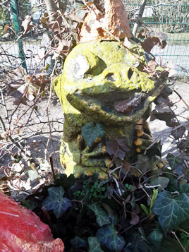 Eine wunderliche Gartenstatue in Form einer lächelnden Kreatur in Säulenform, umschlungen von Pflanzen mit sichtbarem Laub im Hintergrund.