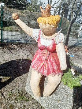 Eine verwitterte Gartenstatue einer weiblichen Figur in einem roten Kleid, die ein Zepter hält und eine Krone auf dem Kopf trägt und in einem sonnigen Außenbereich mit spärlicher Vegetation steht.