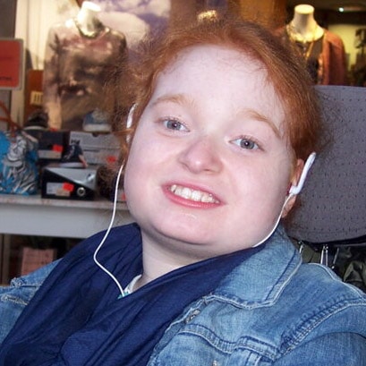Eine junge Frau mit roten Haaren und Kopfhörern lächelt in die Kamera, sie trägt eine Jeansjacke und einen blauen Schal.