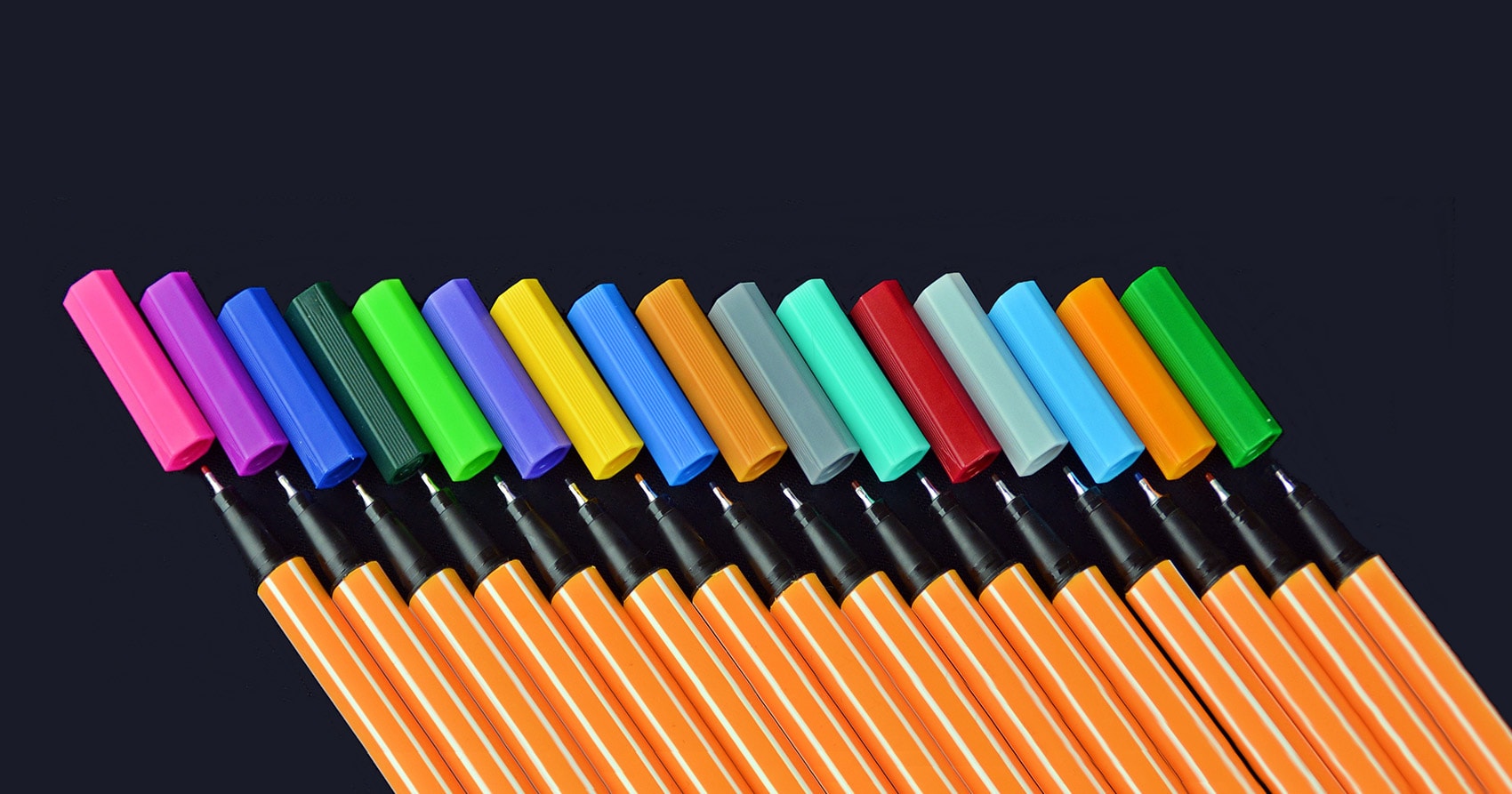 Seitenbild-Konkretisierungen: Exemplarische Planungen: Eine Reihe orangefarbener Stifte mit verschiedenfarbigen Kappen, darunter Rosa, Lila, Blau, Grün, Gelb und Rot, diagonal angeordnet vor einem dunklen Hintergrund.