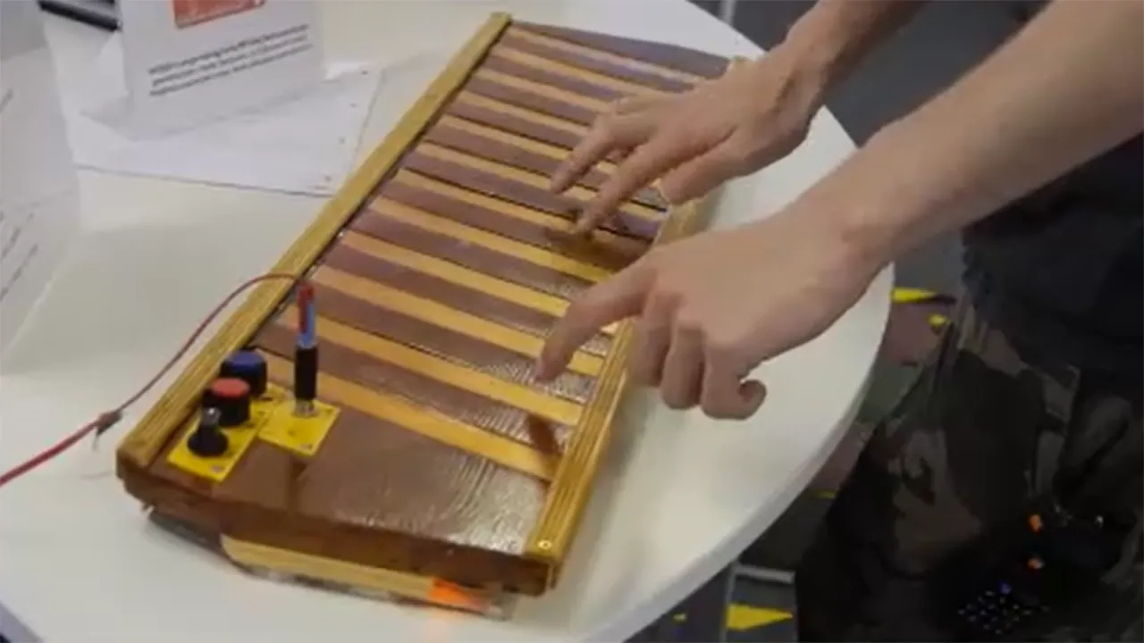 Zwei Hände spielen auf einem weißen Tisch ein handgefertigtes elektronisches Instrument mit Holztasten.