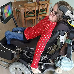 Ein jungesFrau in einem motorisierten Rollstuhl interagiert mit einem Touchscreen-Gerät. Sie trägt ein rot gepunktetes Hemd und Jeans.