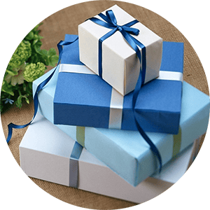 Drei hübsch verpackte Geschenkboxen sind gestapelt. Die obere Box ist weiß mit einer dunkelblauen Schleife, die mittlere ist dunkelblau mit einer weißen Schleife und die untere Box ist hellblau mit einer hellblauen Schleife. Im Hintergrund ist teilweise eine grüne Pflanze zu sehen.