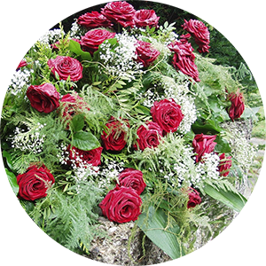 Ein kreisförmiges Blumenarrangement aus zahlreichen roten Rosen und verschiedenen grünen Blättern, dazwischen kleine weiße Blüten, vermutlich Schleierkraut. Die Blumen sind üppig und dicht angeordnet.
