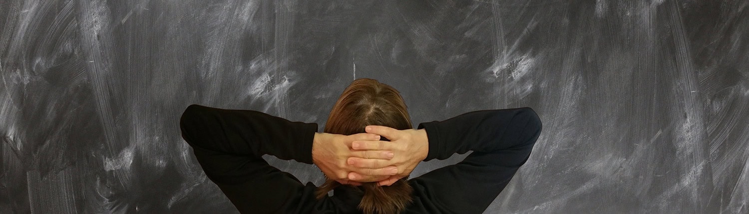Seitenbild-Orientierungsplan: Eine Person mit langen Haaren steht von hinten gesehen mit verschränkten Händen hinter dem Kopf. Sie blickt auf eine dunkle Tafel mit schwachen Kreidemarkierungen. Die Person trägt ein schwarzes langärmeliges Hemd.