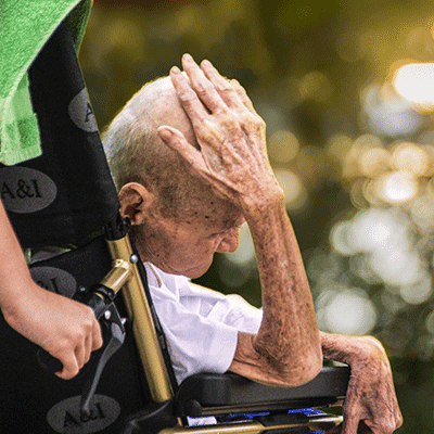 Eine ältere Person mit Glatze sitzt in einem Rollstuhl und stützt ihren Kopf auf ihre Hand. Eine Person steht daneben und hält den Griff des Rollstuhls.
