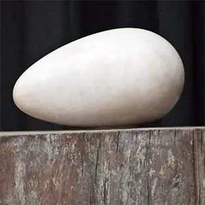 Glatte, weiße, eiförmige Steinskulptur auf einer verwitterten Holzoberfläche mit dunklem Hintergrund.