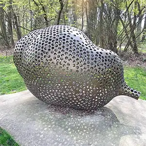 Eine große, perforierte Metallskulptur in Form einer abstrakten Samenkapsel steht auf einem Betonsockel in einem Waldgebiet.